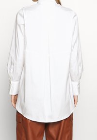 Camicia bianca a maniche lunghe con colletto alto, dettaglio di piega sul retro e orlo arrotondato. Realizzata in tessuto liscio, con polsini abbottonati.
