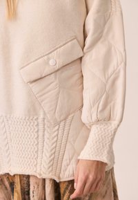 Suéter de crema acolchado con un fondo de punto texturizado, que cuenta con un bolsillo con solapa y puños acanalados. Fabricado con un tejido suave y un diseño en capas.