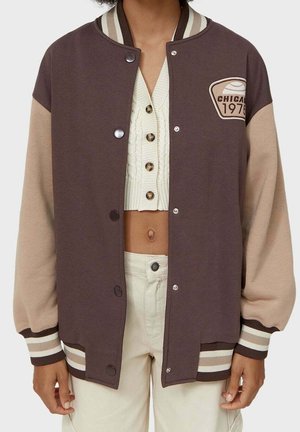 Persoon draagt een bruine en beige varsityjas met "Chicago 1975" badge, crèmekleurig cropped gebreid topje en hoge taille crèmekleurige broek.