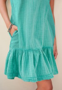 Een lichtteal, textuurvolle jurk met een gerimpelde zoom en een zijzak. De stof heeft een gekreukeld patroon, wat een zachte val geeft.