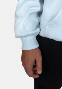 Felpa azzurra chiara con polsini a coste; presenta un logo a diamante ricamato bianco sulla manica; texture morbida, design casual.