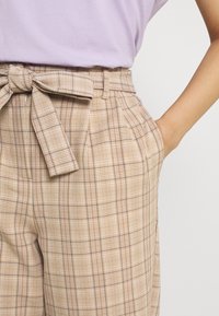 Kvinna i beiga rutiga högmidjade byxor med ett knytbälte och handen i fickan, kombinerat med en lavendelfärgad topp.