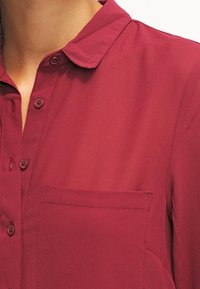 Burgundy button-up shirt with a collar, featuring a single chest pocket and textured fabric. Buttons are round and match the shirt color.