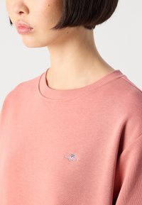 Close-up of a person wearing a pink crewneck sweatshirt with a small GANT logo on the chest.