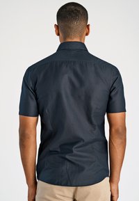 Man staat met zijn rug naar de camera, gekleed in een aansluitend, kort mouw donkerblauw shirt en lichtbeige broek, tegen een eenvoudige witte achtergrond.