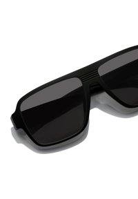 Mustad ristkülikukujulised päikeseprillid tumedate läätsedega. Raamil on matt viimistlus ja õrn tekstuur käepidemetel.