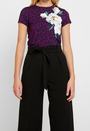 Kobieta w fioletowej koszuli w panterkę z białymi kwiatowymi detalami oraz czarnych, wysokich spodniach z paskiem wiązanym w talii.