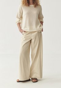 Cremfarbener Strickpullover mit lockerem Schnitt und geripptem Halsausschnitt, kombiniert mit weiten beigen Hosen und braunen flachen Sandalen.