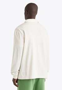 Camicia a maniche lunghe di colore crema con una vestibilità rilassata, colletto a bottoni e tessuto strutturato. Presenta un piccolo etichetta in basso a sinistra.