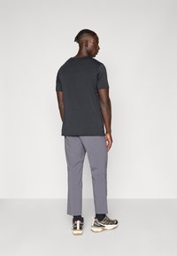 Black short-sleeve t-shirt paired with light grey trousers; model wearing black and beige athletic shoes; rear view.