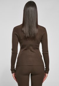 Brun ribbet langermet topp med tettsittende design, rund hals og smale mansjetter, sammen med matchende ribbet bukser, sett fra bak.