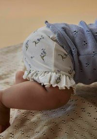 Medvilninės kūdikių šortai su baltai mėlynu gėlių raštu ir šoniniu volanu. Iš dalies matomas mėlynas megztas viršus.