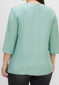 Ljus teal-blus med tre fjärdedelsärmar, slät yta, rundad halsringning och avslappnad passform, sedd bakifrån.