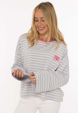Junge Frau lächelt und schaut nach unten, trägt ein blau-weiß gestreiftes langärmeliges Shirt mit einem pinken Fleck und weiße Hosen.