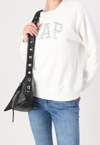 Borsa in pelle nera con forma triangolare e dettagli in metallo argentato. La tracolla presenta accenti circolari. Indossata con un maglione bianco e jeans blu.