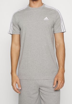 Hombre vistiendo una camiseta Adidas de color gris claro con tres rayas blancas en los hombros y el logo de Adidas en el pecho izquierdo.