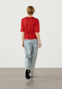 Camicia rossa a maniche corte con una vestibilità rilassata, abbinata a jeans azzurri chiari e larghi e scarpe slip-on nere. Vista posteriore.