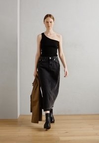 Black one-shoulder top, high-waisted black denim skirt, brown outerwear draped, and black ankle boots. Plain background with light wood floor.