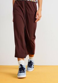 Calças desportivas castanhas com riscas laterais cor de rosa, feitas de um tecido suave. Combinadas com ténis azul claro e meias brancas.