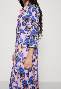 Kwiatowa sukienka z różowym tłem, dużymi niebieskimi i pomarańczowymi kwiatami, długimi rękawami, zapięciem na guziki oraz zwiewnym krojem.