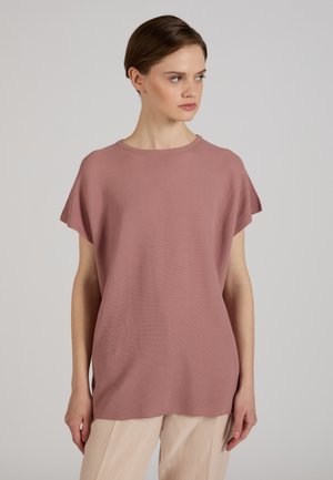 Femme portant un haut ample rose poudré à manches courtes et un pantalon beige clair, regardant vers sa droite sur un fond uni.