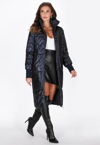 Lang marineblå pufferfrakke med høj krave, lynlås-lukning og ribbede cuffs. Har et design der går til skinnebenene med en blank overflade og en subtil syet mønster.