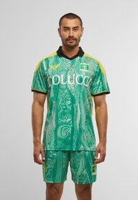 Mann in grünem gemustertem Fußballtrikot mit gelben Akzenten, schwarzem Kragen und Logos auf Brust und Shorts, steht vor einem schlichten Hintergrund.