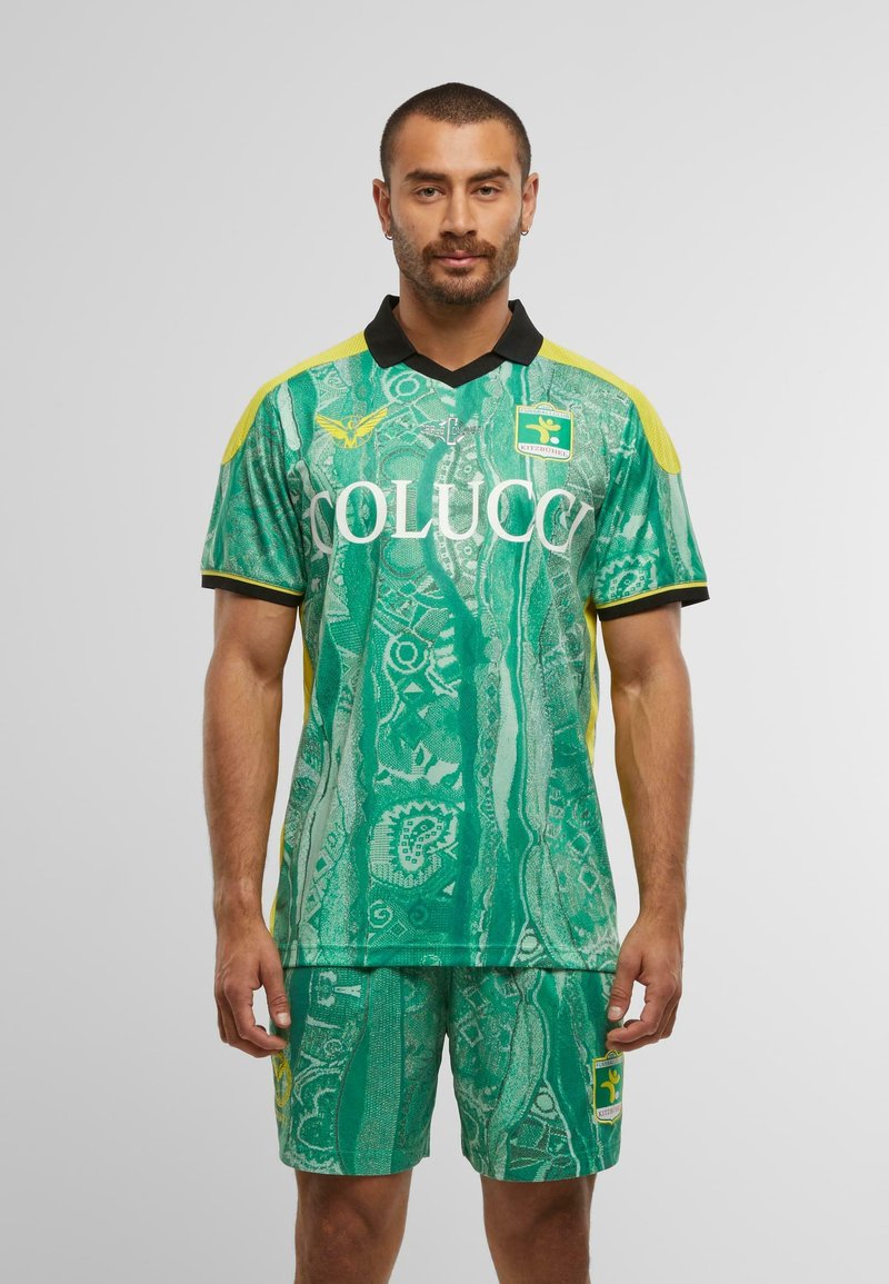 Mann in grünem gemustertem Fußballtrikot mit gelben Akzenten, schwarzem Kragen und Logos auf Brust und Shorts, steht vor einem schlichten Hintergrund.