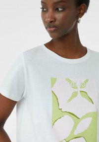 T-shirt blanc à manches courtes avec un imprimé graphique vert et rose de feuilles, portant le mot "vacances" en texte clair, fabriqué en tissu doux.