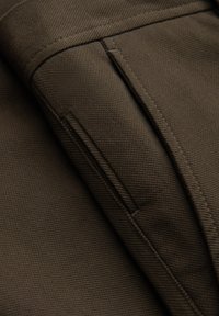Tessuto marrone con una trama testurizzata, caratterizzato da un dettaglio con tasca e cuciture pulite lungo i bordi, che evidenziano il suo design strutturato.