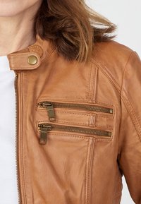 Bruine leren jack met een opstaande kraag, twee voorzakken met rits en bronzen hardware, en subtiele stiksels op de schouders.