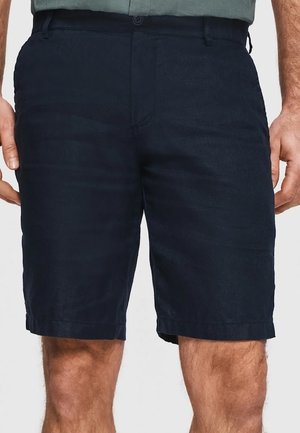 Mężczyzna w ciemnoniebieskich krótkich spodenkach do kolan z przednimi kieszeniami i zapinanym na guziki paskiem, stojący z rękami luźno opuszczonymi wzdłuż ciała.