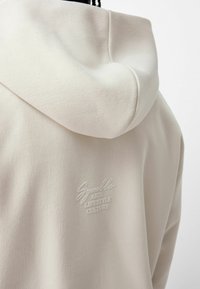 Sudadera color beige claro con un tejido texturizado, una capucha grande y un logo bordado en la parte trasera. Diseño simple y contemporáneo.