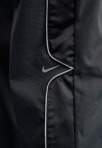 Černé sportovní kalhoty s hladkou, matnou texturou. Obsahují stříbrné logo Nike a bílé lemování podél švů.