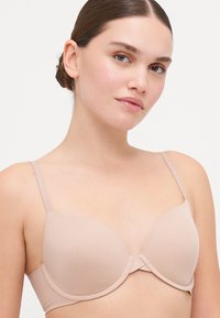 Beige padded bra with adjustable straps, smooth texture, and a seamless design. Decorative center panel for added detail.
