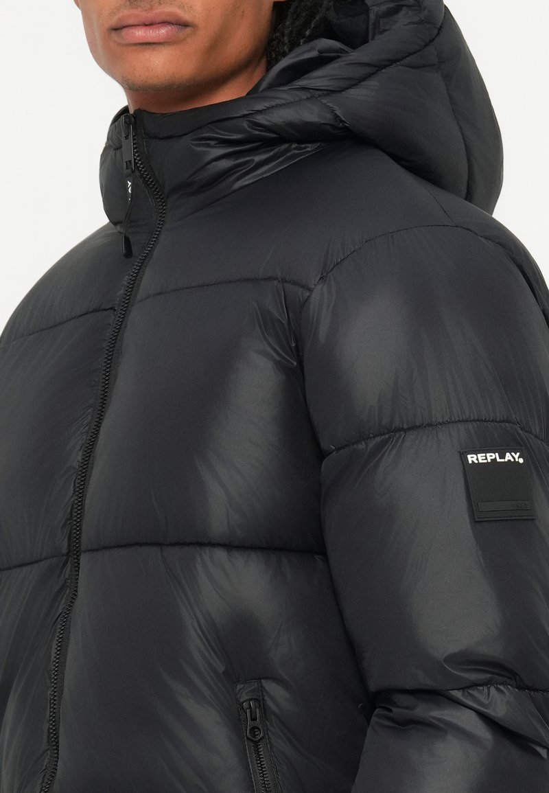 Veste rembourrée noire avec un col haut, fermeture éclair et un patch logo sur le bras. Elle présente une texture lisse et brillante ainsi que deux poches latérales.