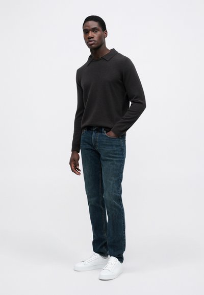 Fiatal férfi áll, az egyik keze a sötét színű galléros pulóverében, kék farmerben és fehér sportcipőben egy sima háttérrel.