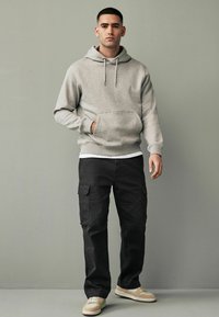 Sudadera gris con capucha y bolsillo tipo canguro, combinada con pantalones cargo negros con bolsillos laterales y zapatillas beige. Tejido texturizado.