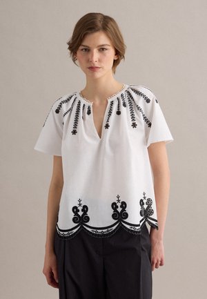 Giovane donna che indossa una camicetta bianca con ricamo floreale nero e maniche corte, abbinata a pantaloni neri, in piedi davanti a uno sfondo uniforme.