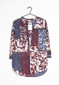 Blouse à manches longues avec motifs patchwork floraux et géométriques multicolores en bordeaux, bleu, blanc et pêche, suspendue sur un cintre noir.