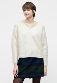 Lys kremfarget cardigan med V-hals, teksturert strikk, og fem knappelukkinger, kombinert med et mørkt rutete skjørt med blå og grønne nyanser.