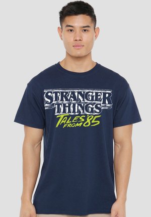 Junger Mann trägt ein dunkelblaues T-Shirt mit dem Aufdruck „Stranger Things Tales from 85“ und steht vor einem einfarbigen hellgrauen Hintergrund.
