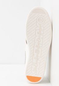 Suela de zapatilla deportiva con material de goma blanca, agarre texturizado, logotipo "SUPERDRY" en relieve y un detalle naranja en el talón.