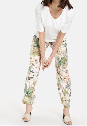 Mujer con blusa blanca de manga corta y escote en V, pantalones con estampado floral y sandalias beige de tacón, de pie con las manos juntas al frente.