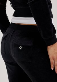 Black velour pants feature a fitted design with rear pockets, accented by a silver grommet detail. Soft texture, elastic waistband visible.