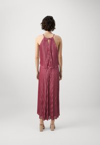 Robe plissée en rose profond, sans manches avec un détail noué à l'arrière, présentant une silhouette fluide et un ourlet au niveau des chevilles. Texture lisse, tissu doux.