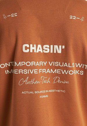 Tangerine katoenen T-shirt met witte bedrukte tekst en zinnen zoals "CHASIN'" en "Authen-Tech Denim," tegen een soepele stoftextuur.