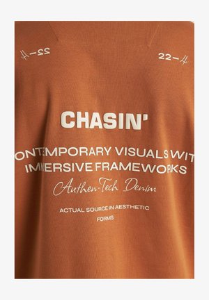 Orange bomull t-shirt med vit tryckt text med fraser som "CHASIN'" och "Authen-Tech Denim," mot en slät tygstruktur.