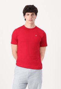 Camiseta corta roja de algodón, con cuello redondo y un pequeño logo en el pecho. Combinada con pantalones de color claro.