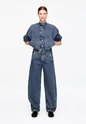 Mujer de pie, vistiendo una chaqueta vaquera azul oscuro de corte holgado con mangas abullonadas y vaqueros anchos a juego, con las manos en los bolsillos y zapatos negros.